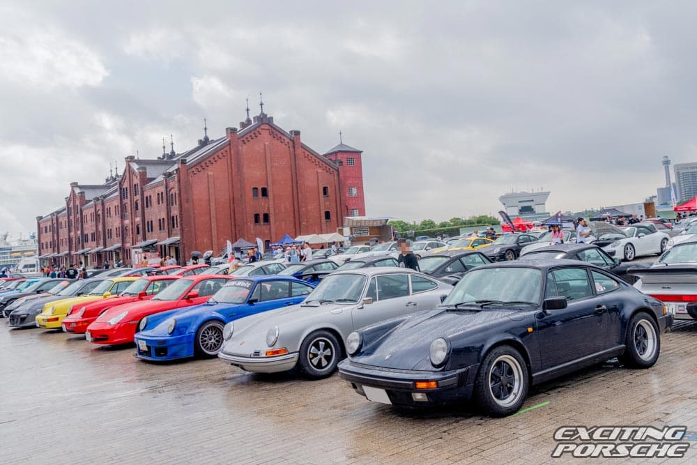 2024 年 EXCITING PORSCHE(横浜 赤レンガ倉庫)で、雨上がりの会場に色とりどりの参加者のポルシェが並ぶ様子